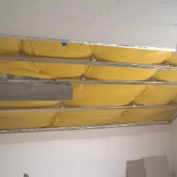 Sufit podwieszany w trakcie montażu: metalowa konstrukcja, żółta izolacja. Widoczny fragment wentylacji i przewód. Białe ściany, surowy stan wykończenia.