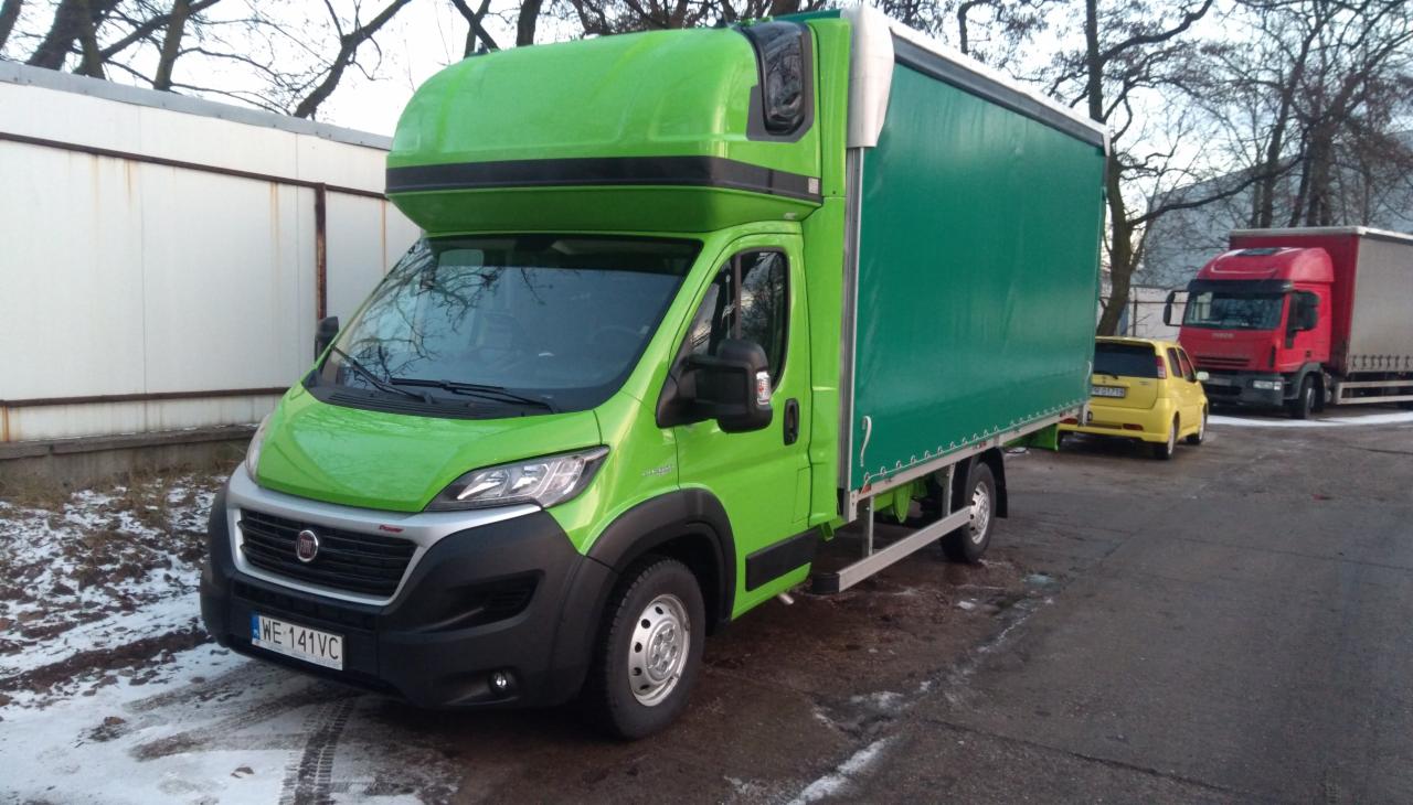 Zielony samochód dostawczy Fiat Ducato z plandeką, zaparkowany na placu, z widoczną tablicą rejestracyjną WE 141VC, w tle żółty samochód osobowy i czerwona ciężarówka.