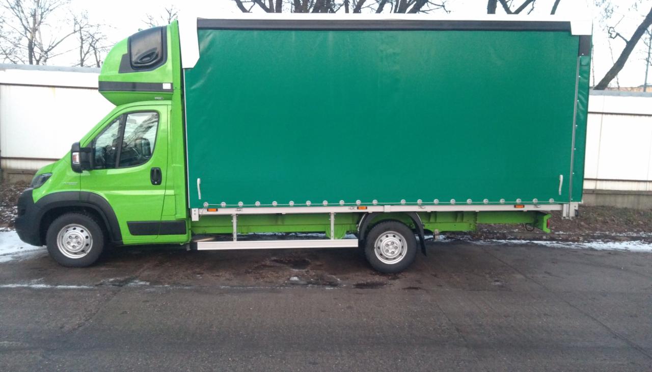 Zielony samochód dostawczy z plandeką, zaparkowany na asfalcie, z widocznymi drzewami w tle.