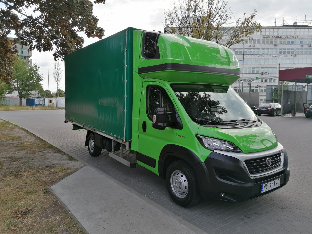 Zielony samochód dostawczy z plandeką na parkingu miejskim, widok z boku, tablica rejestracyjna WE 141VC.