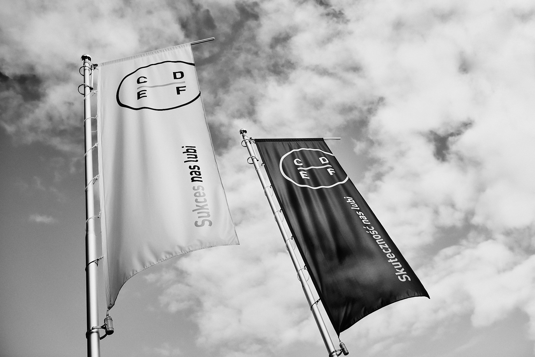 Dwa flagi na tle pochmurnego nieba: jedna biała z napisem 'Sukces nas lubi' i logo CDF, druga czarna z napisem 'Skuteczność nas lubi' i logo CDF.