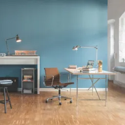 Nowoczesne biuro domowe z błękitną ścianą, drewnianą podłogą i metalowymi akcentami. Biurko z laptopem pod lampką, krzesło obrotowe, okno z grubym murem.