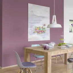 Fioletowa ściana w jadalni z drewnianym stołem, nowoczesną lampą i abstrakcyjnym obrazem. Jasna podłoga i krzesła w pastelowych kolorach dopełniają wnętrze.