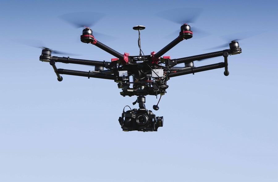 Czarny dron z kamerą Lumix w locie na tle błękitnego nieba. Widoczne wirniki i elementy konstrukcyjne drona. Sprzęt do filmowania z powietrza.