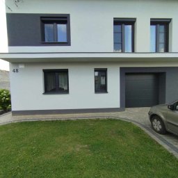 P.P.H.U EKO-PLAST JACEK BADORA - Nowoczesny dom dwupiętrowy w odcieniach szarości z ciemnymi oknami PCV z roletami zewnętrznymi, bramą garażową segmentową i zadbanym trawnikiem, widok z poziomu trawnika.