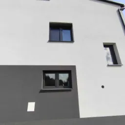 Nowoczesna elewacja budynku z trzema oknami o ciemnych ramach, dwa z nich prostokątne, jedno wąskie, pionowe, na tle białej i szarej ściany.