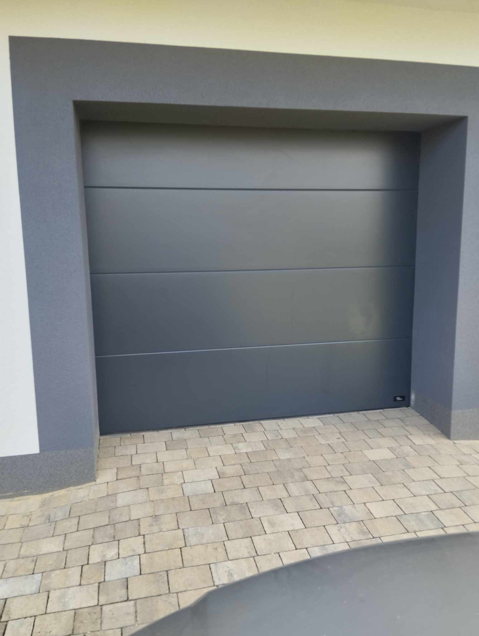 Zamknięta, segmentowa brama garażowa w kolorze grafitowym, z widocznymi poziomymi panelami, osadzona w szarej ramie, na tle kostki brukowej.