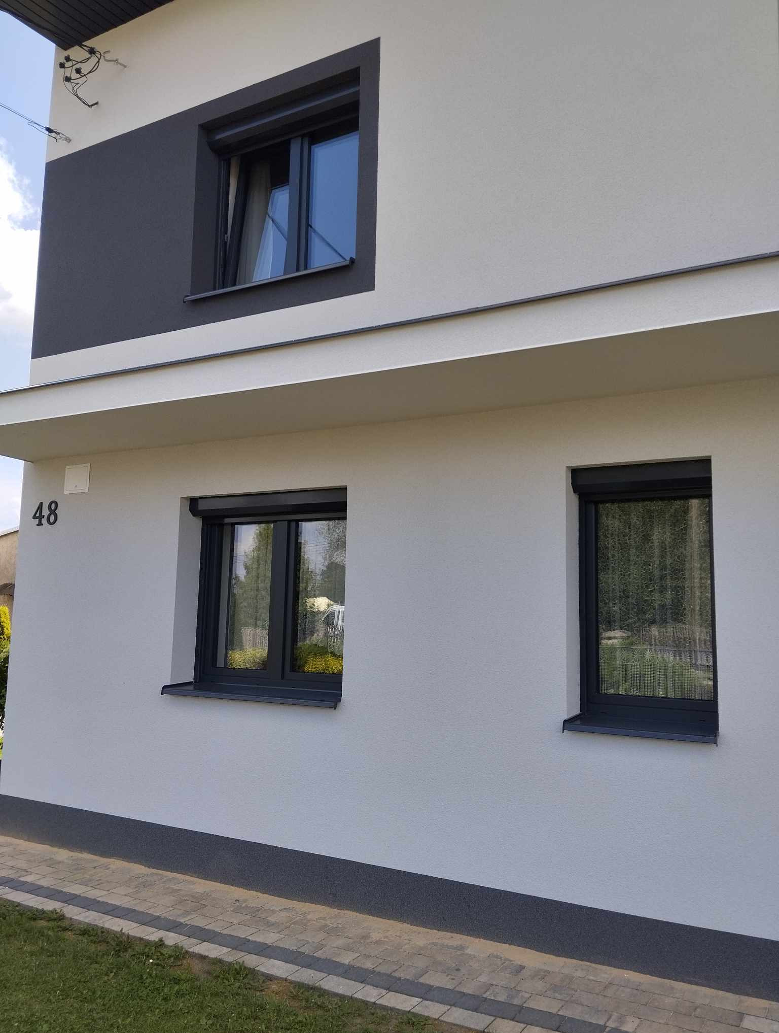 Elewacja domu z trzema oknami z roletami zewnętrznymi w kolorze antracytowym, widok z poziomu chodnika, numer domu '48' po lewej stronie.