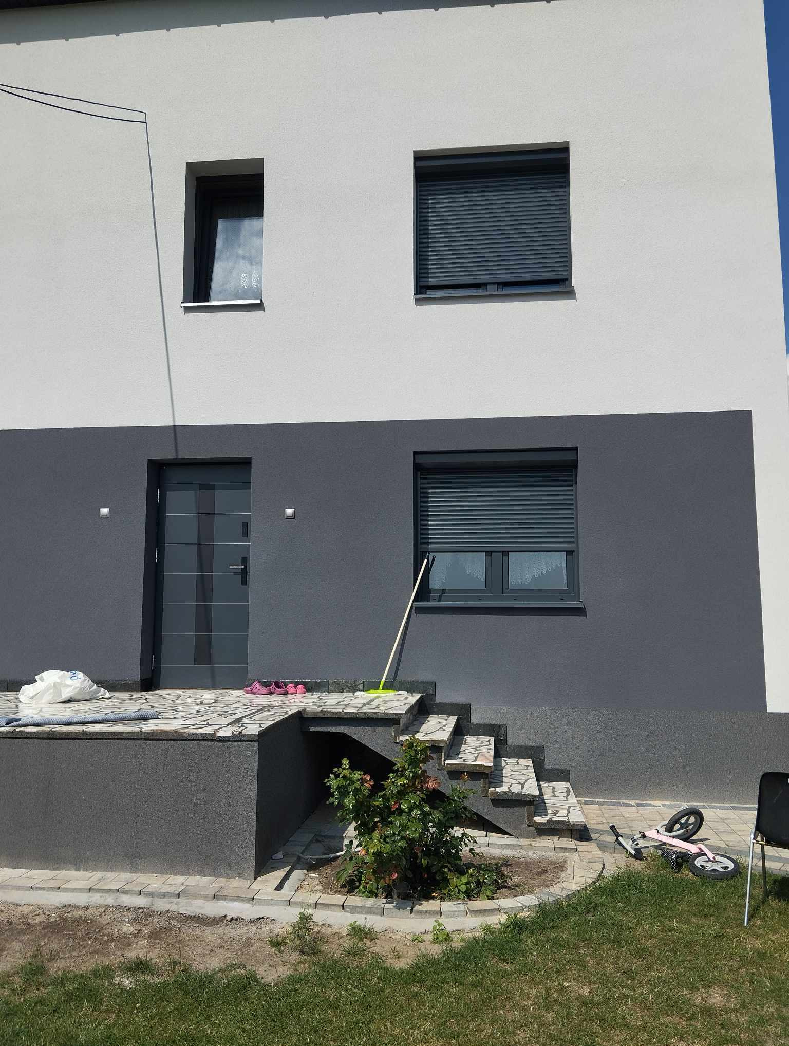Elewacja domu jednorodzinnego z szarymi oknami z roletami zewnętrznymi, szare drzwi wejściowe, schody z kostki brukowej i rozrzucone przedmioty na podwórku.