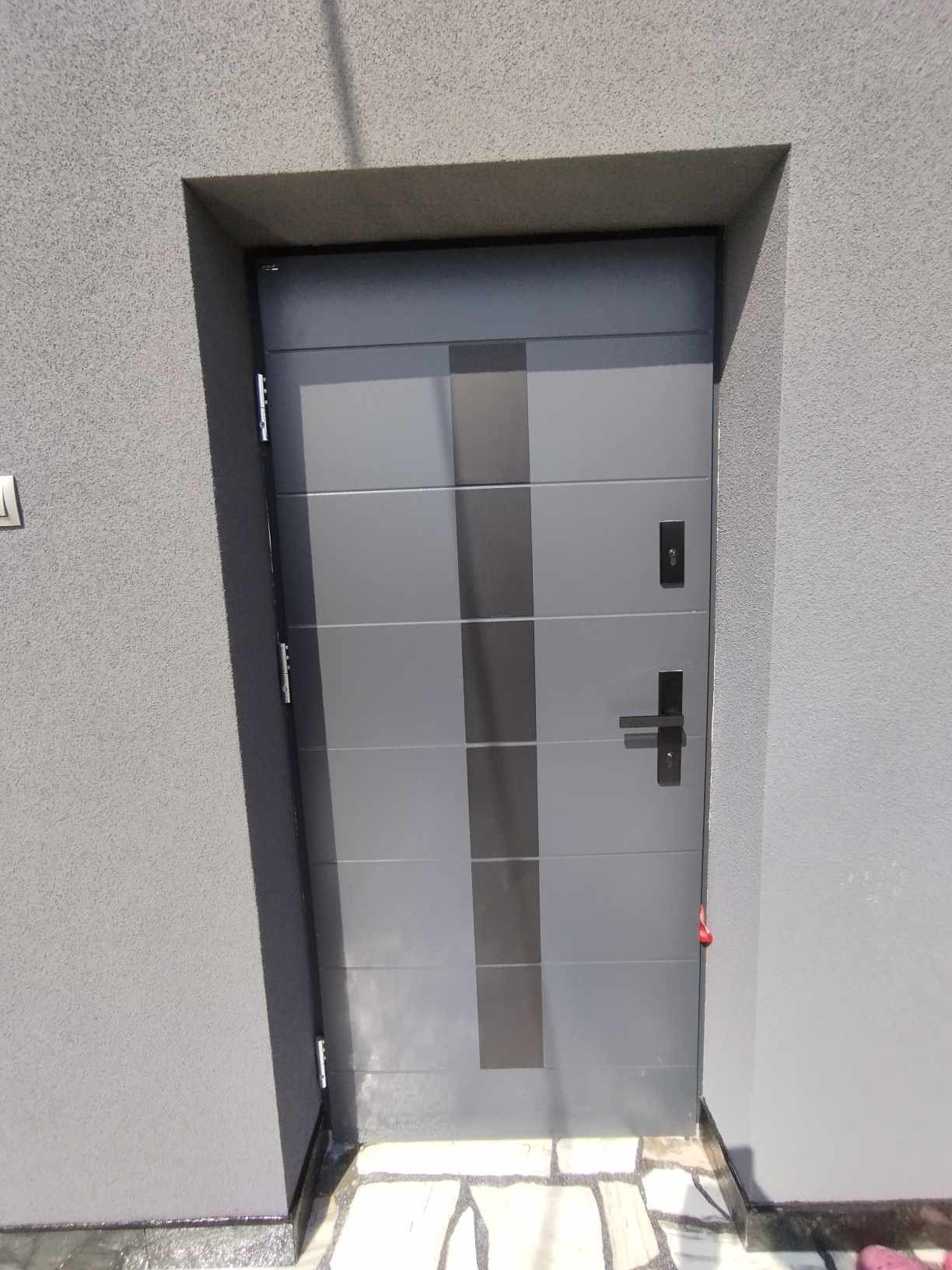 Szare, nowoczesne drzwi wejściowe z czarnym, pionowym pasem, zamontowane w szarej elewacji, widoczne zawiasy i klamka.