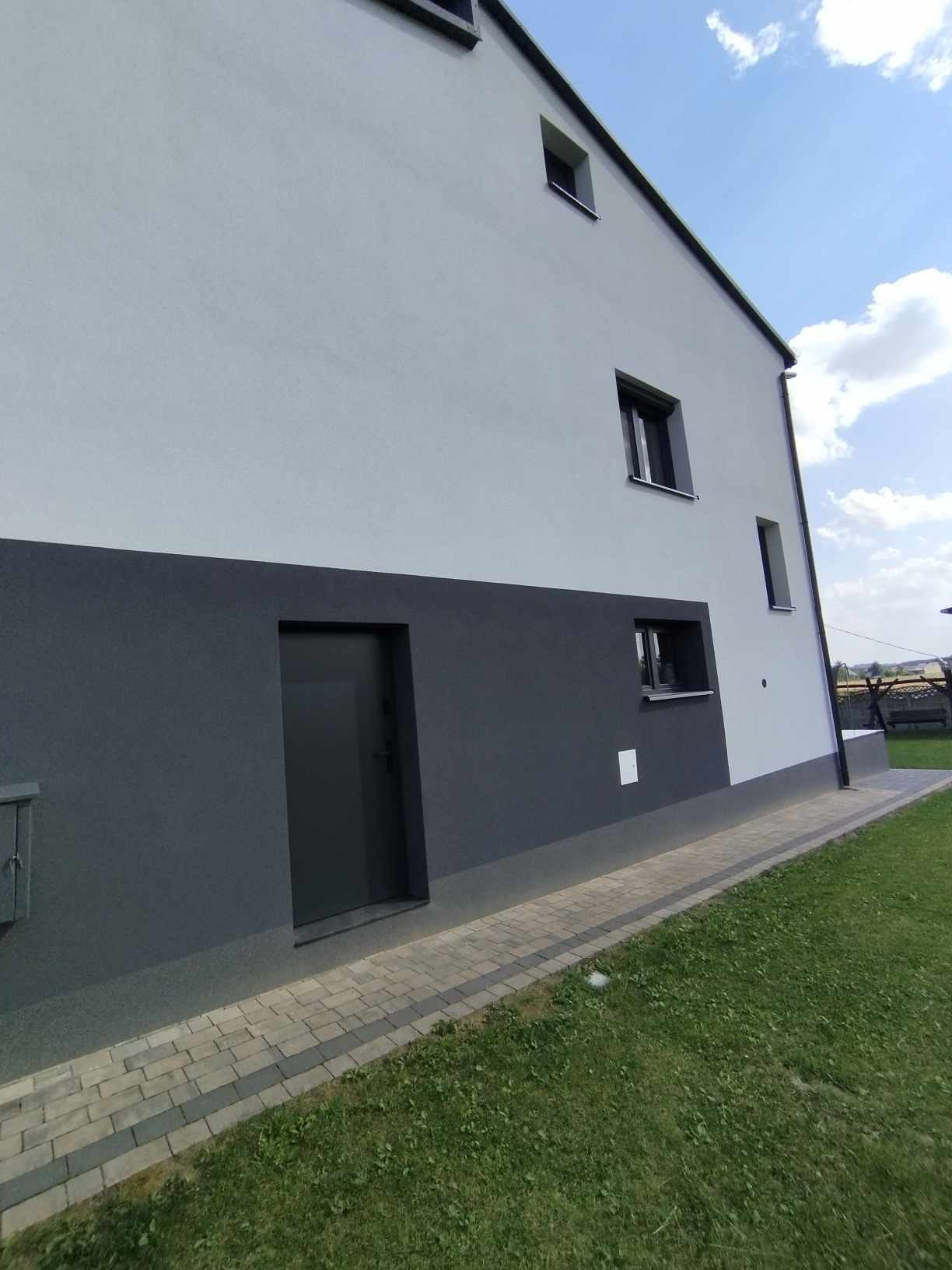 Elewacja budynku dwukolorowa, szaro-biała, z małymi oknami i ciemnymi drzwiami wejściowymi, widok z poziomu trawnika.
