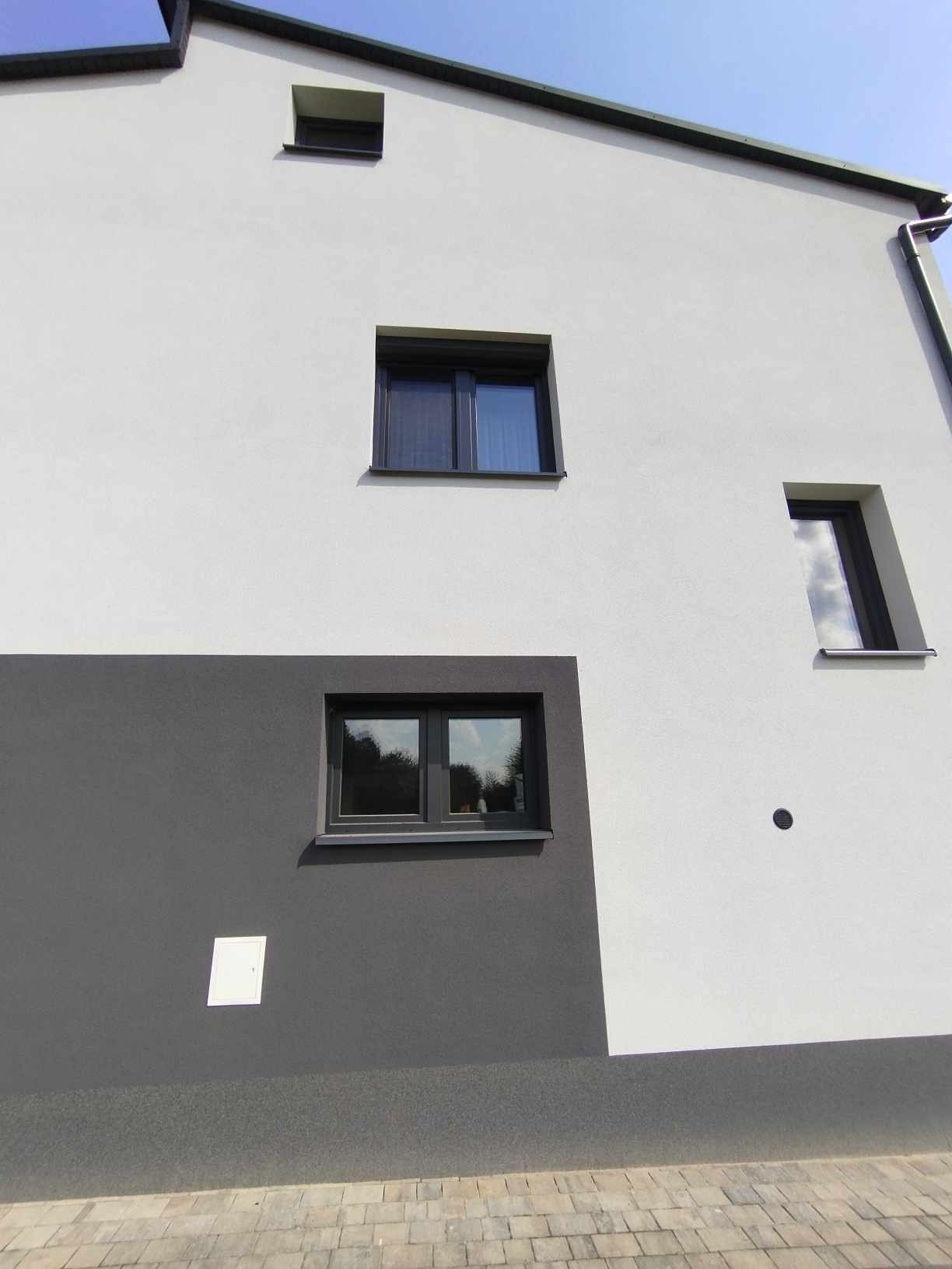 Nowoczesna elewacja budynku z trzema oknami o ciemnych ramach, dwa z nich prostokątne, jedno wąskie, pionowe, na tle białej i szarej ściany.