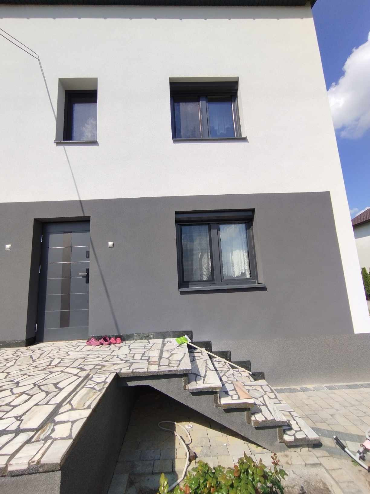 Dwukondygnacyjny dom z szarą stolarką okienną i drzwiami wejściowymi, jasna elewacja górnej kondygnacji kontrastuje z szarą dolną, kamienne schody prowadzą do drzwi.