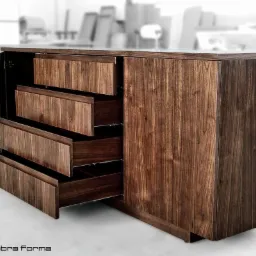 Nowoczesna szafka z ciemnego drewna z wysuwanymi szufladami, widoczne metalowe prowadnice, logo firmy w lewym dolnym rogu.