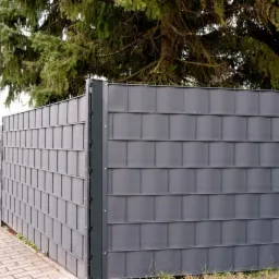 Grafitowe ogrodzenie panelowe z widocznymi słupkami, na tle zieleni drzew, z perspektywy chodnika z kostki.