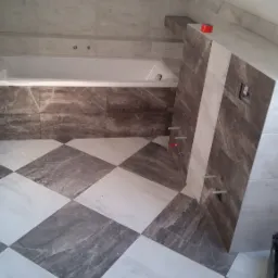 Ukończona łazienka na poddaszu z biało-szarą szachownicą na podłodze, marmurowe płytki na ścianach, zabudowana wanna i fragment ściany z wystającymi rurami instalacji wodnej, zabezpieczona...