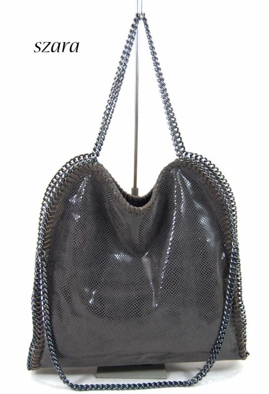 Szara torebka typu shopper z łańcuszkami na białym tle, zawieszona na metalowym stojaku.