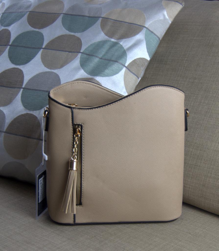 Beżowa, prosta torebka damska z frędzlem przy suwaku kieszeni bocznej, umieszczona na sofie z poduszkami w tle.
