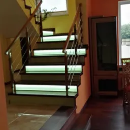 Nowoczesne schody z podświetlanymi stopniami i balustradą z drewnianymi poręczami i nierdzewnymi linkami, widok z dołu na jasne, oświetlone wnętrze.