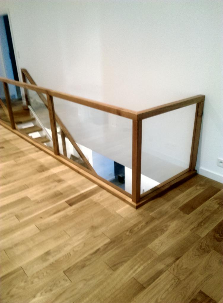 Fragment drewnianej balustrady ze szklanym wypełnieniem, zabezpieczającej schody, widoczna z perspektywy podłogi z parkietu.