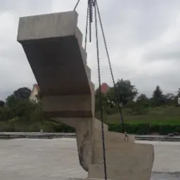 Prefabrykowane betonowe schody wiszące na łańcuchach z dźwigu na placu budowy, widoczne budynki w tle.