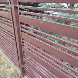 SPAWMET - Fragment brązowego ogrodzenia panelowego z poziomych listew, widok z boku na tle zielonych iglaków.