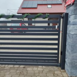 SPAWMET - Nowoczesna brama wjazdowa z poziomymi panelami w kolorze grafitowym, ozdobiona świątecznym łańcuchem, na tle domu z czerwoną dachówką i panelami słonecznymi.