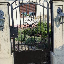 SPAWMET - Czarna, metalowa brama z ozdobnymi elementami, umieszczona między dwoma kamiennymi słupkami z lampami, prowadząca do ogrodu z widocznym domem w tle.