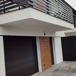 SPAWMET - Nowoczesny dom z czarnymi bramami rolowanymi, drewnianą okładziną i poziomymi czarnymi barierkami balkonowymi, widok z dołu na szarej kostce brukowej.