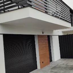 Nowoczesny dom z czarnymi bramami rolowanymi, drewnianą okładziną i poziomymi czarnymi barierkami balkonowymi, widok z dołu na szarej kostce brukowej.