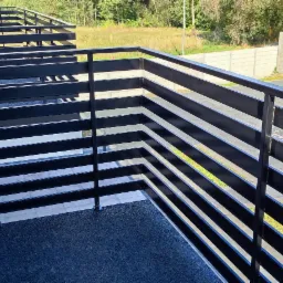 Nowoczesna, czarna balustrada balkonowa z poziomych profili, widok z góry na ciemną, antypoślizgową nawierzchnię balkonu, w tle betonowe ogrodzenie i zieleń drzew.