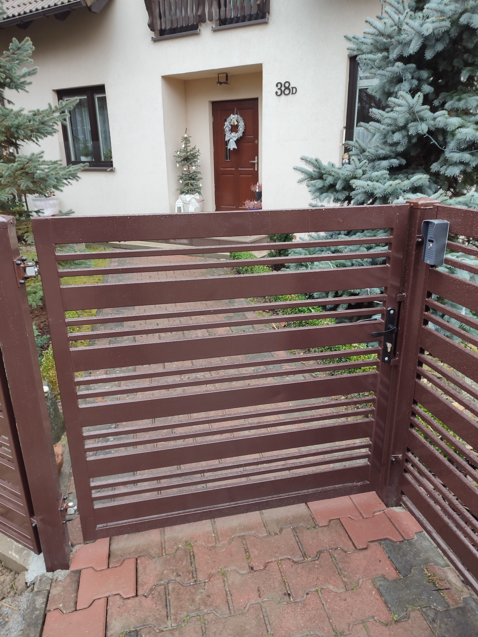 Brązowa, pozioma brama lamelowa z domofonem na słupku, widok na dom z numerem 38D i świątecznym stroikiem na drzwiach.