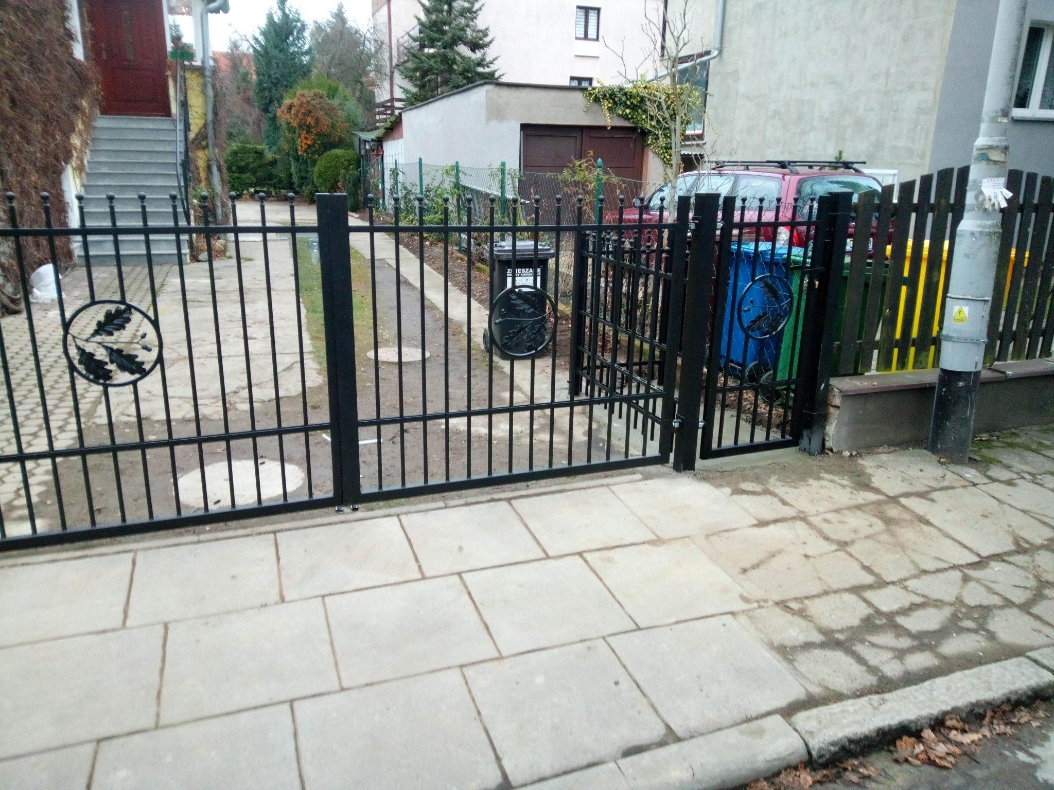 Czarne metalowe ogrodzenie z bramką, ozdobione motywem liści dębu, na tle chodnika z szarych płyt i zabudowań mieszkalnych.