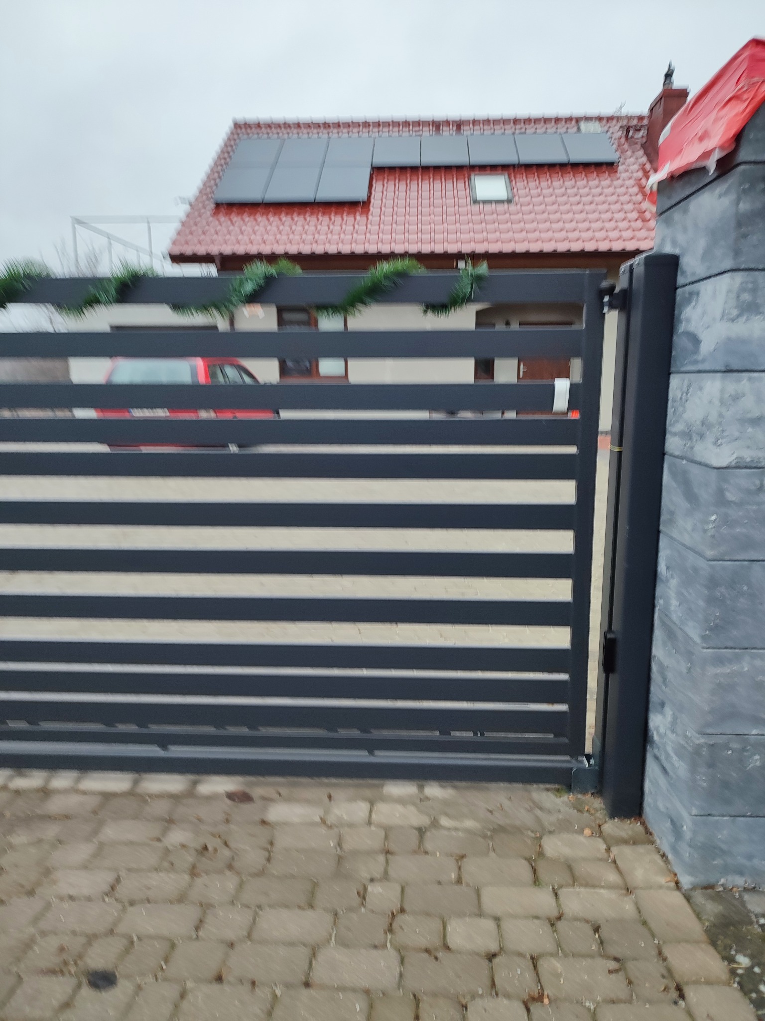 Nowoczesna brama wjazdowa z poziomymi panelami w kolorze grafitowym, ozdobiona świątecznym łańcuchem, na tle domu z czerwoną dachówką i panelami słonecznymi.