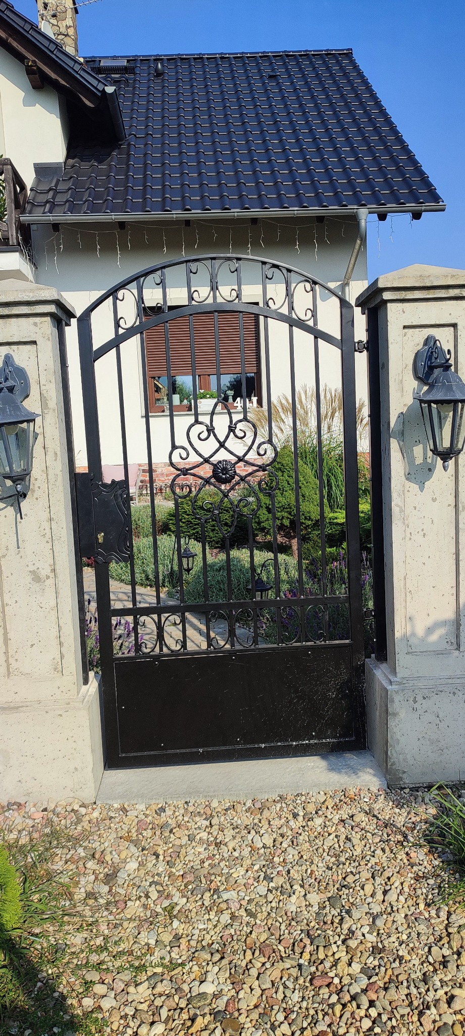Czarna, metalowa brama z ozdobnymi elementami, umieszczona między dwoma kamiennymi słupkami z lampami, prowadząca do ogrodu z widocznym domem w tle.