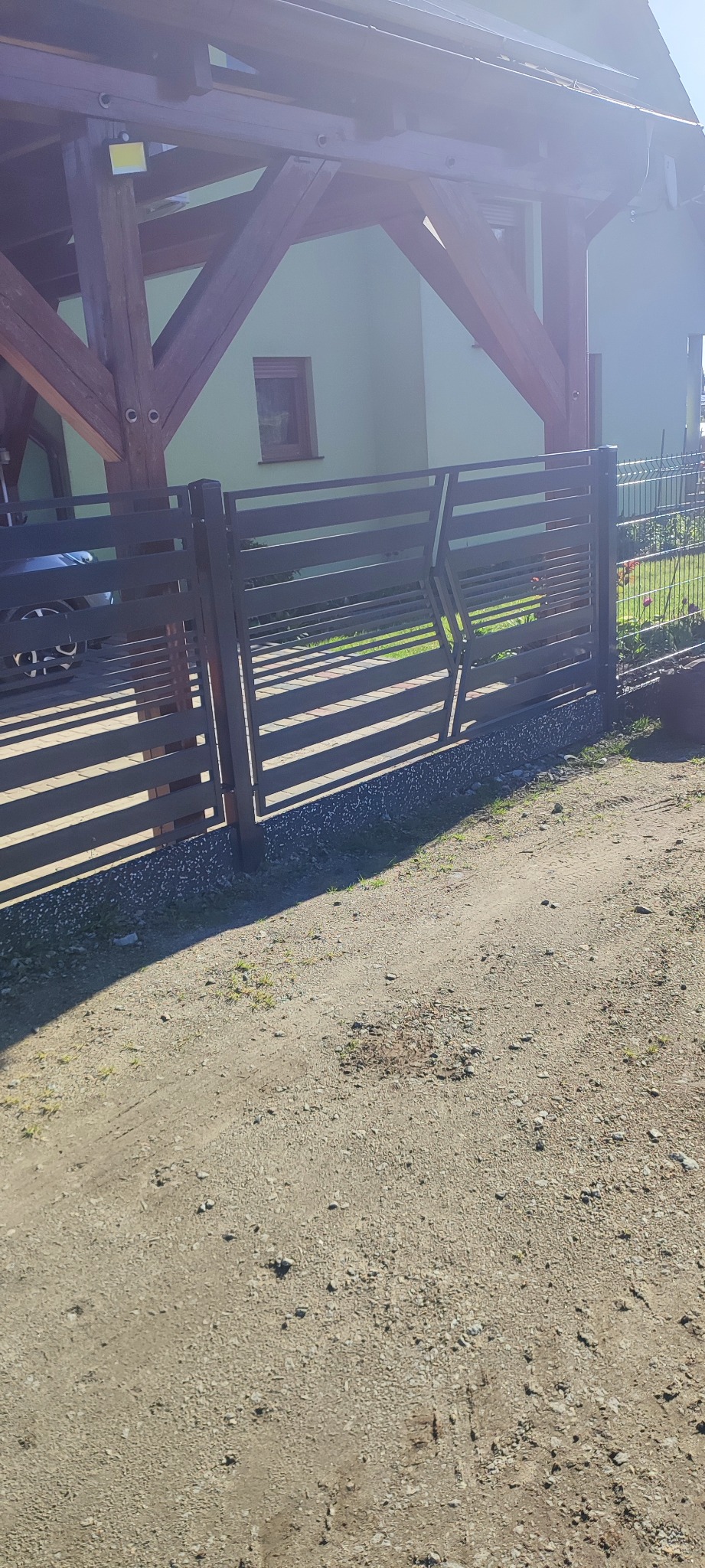 Nowoczesne ogrodzenie metalowe z poziomych profili, z ukośnym elementem dekoracyjnym, widoczne z zewnątrz posesji na tle domu i wiaty.