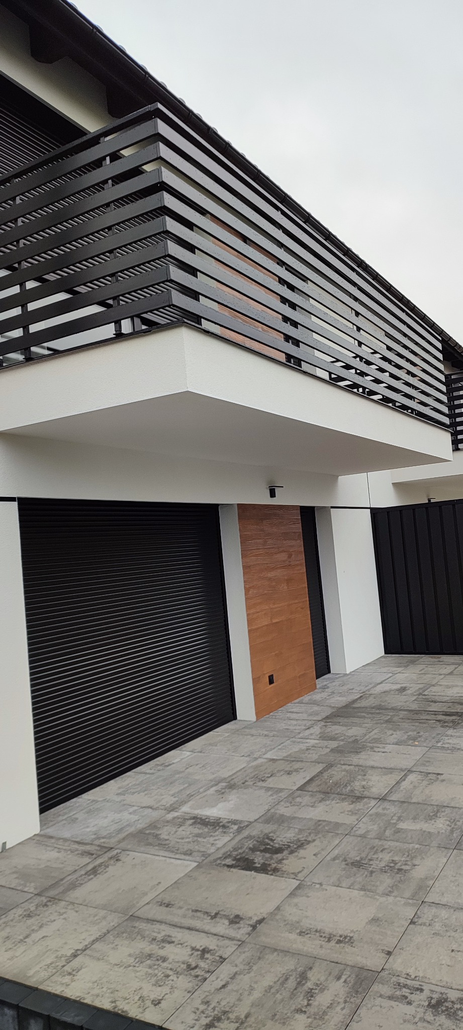 Nowoczesny dom z czarnymi bramami rolowanymi, drewnianą okładziną i poziomymi czarnymi barierkami balkonowymi, widok z dołu na szarej kostce brukowej.