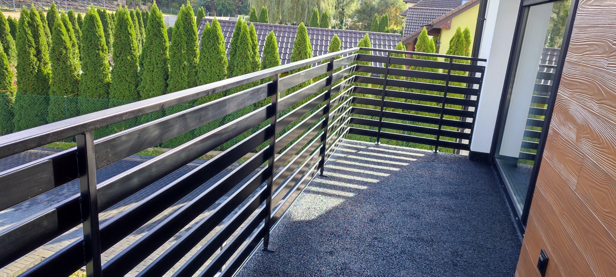 Czarna, pozioma balustrada metalowa na balkonie z widokiem na rząd zielonych tuj i domy w tle. Balustrada z profili zamkniętych, mocowana do podłoża balkonu pokrytego szarym granulatem.