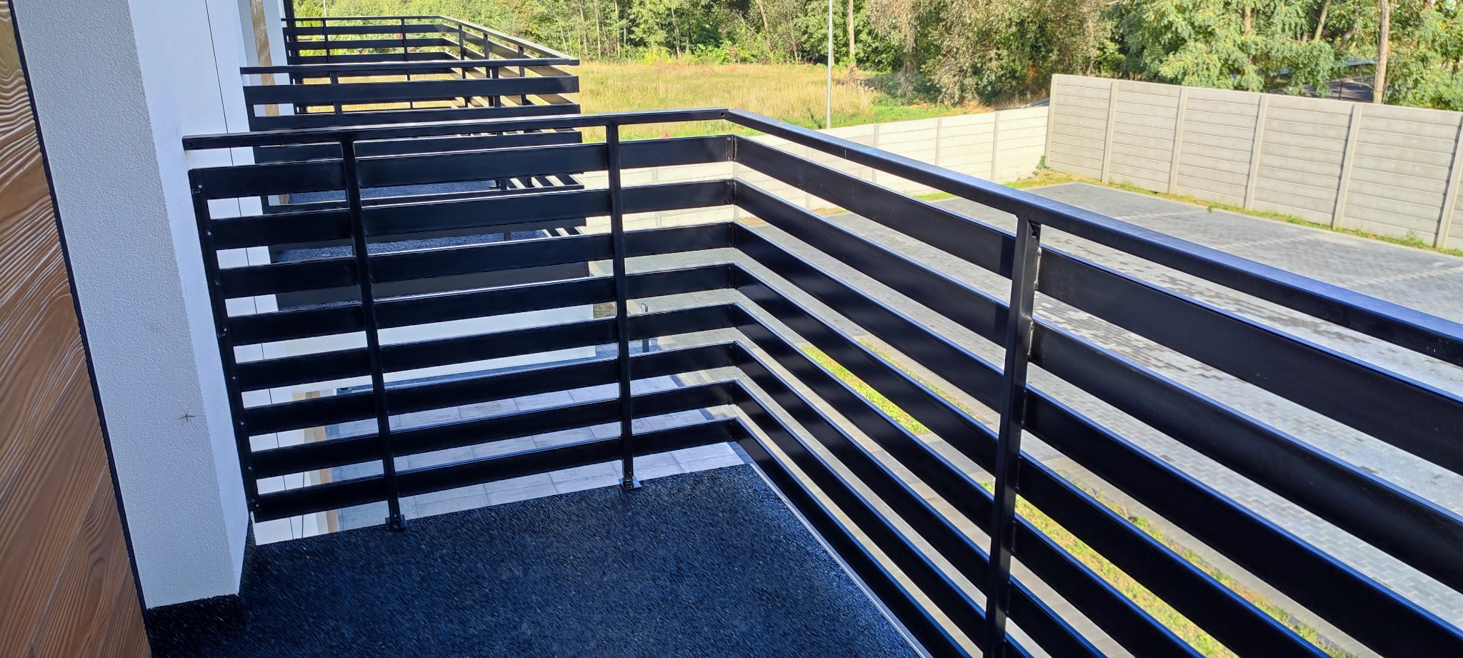 Nowoczesna, czarna balustrada balkonowa z poziomych profili, widok z góry na ciemną, antypoślizgową nawierzchnię balkonu, w tle betonowe ogrodzenie i zieleń drzew.