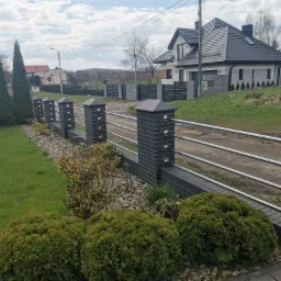 Nowoczesne ogrodzenie posesji z elementami z szarej cegły i metalowymi przęsłami na tle domu z ciemną dachówką i zielonego trawnika.