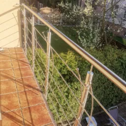 Błyszcząca, metalowa balustrada na balkonie z terakotową podłogą, widok na ogród z kwitnącym drzewem i krzewami, w tle budynek mieszkalny.