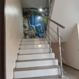 Białe schody z okładziną imitującą drewno, zabezpieczone metalową balustradą z wypełnieniem z cienkich prętów, na ścianie fototapeta z motywem wodospadu.