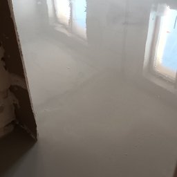 DARKTECH - Świeżo wylana, gładka posadzka samopoziomująca w jasnym odcieniu szarości, odbijająca światło okien. Widoczne surowe ściany w trakcie prac wykończeniowych.