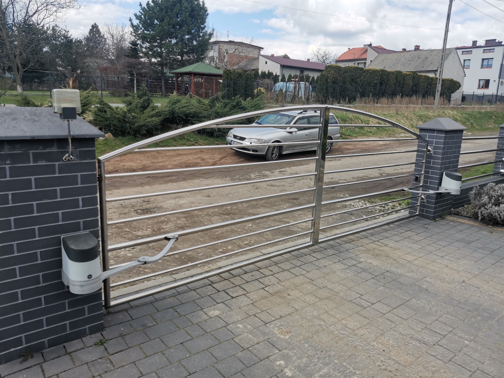 Automatyczna brama wjazdowa z nierdzewnej stali z napędem, widok z zewnątrz na posesję z zaparkowanym samochodem.