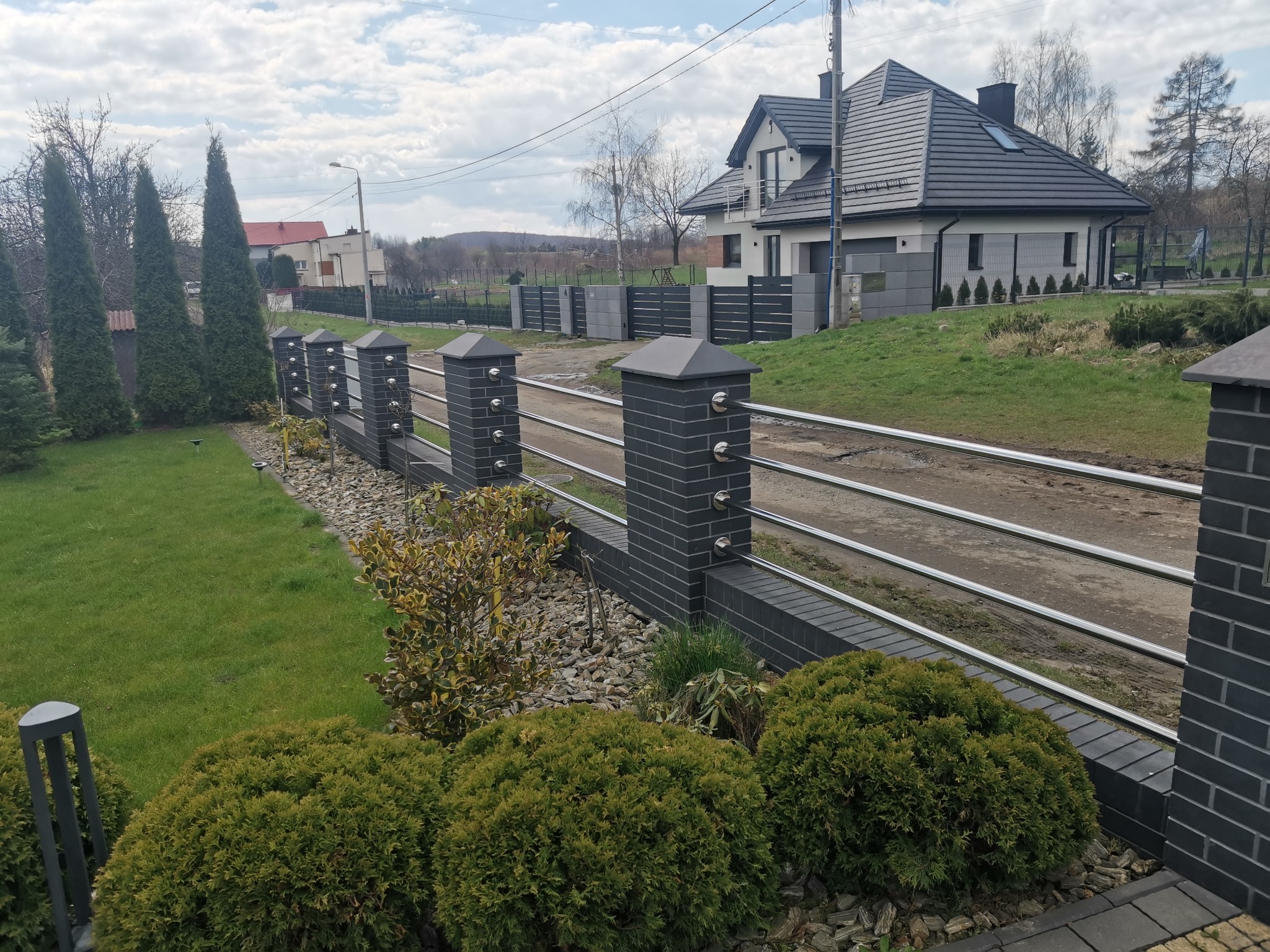 Nowoczesne ogrodzenie posesji z elementami z szarej cegły i metalowymi przęsłami na tle domu z ciemną dachówką i zielonego trawnika.