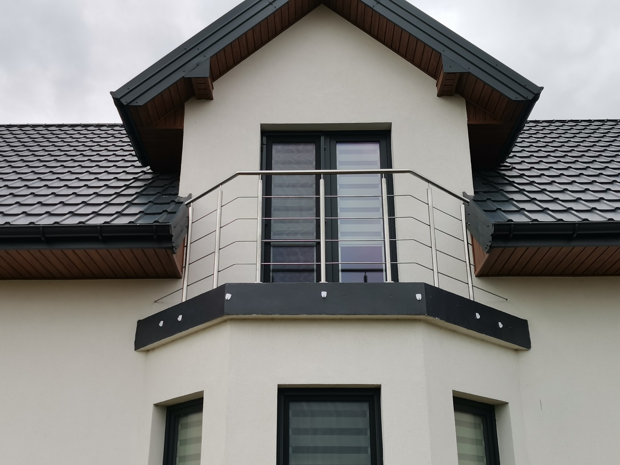 Biały dom z balkonem zabezpieczonym nowoczesną, poziomą, stalową balustradą. Widoczne czarne ramy okienne i dach z ciemnej dachówki.