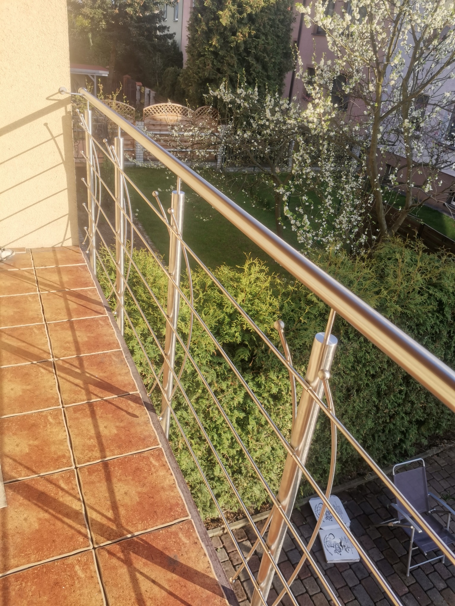 Błyszcząca, metalowa balustrada na balkonie z terakotową podłogą, widok na ogród z kwitnącym drzewem i krzewami, w tle budynek mieszkalny.