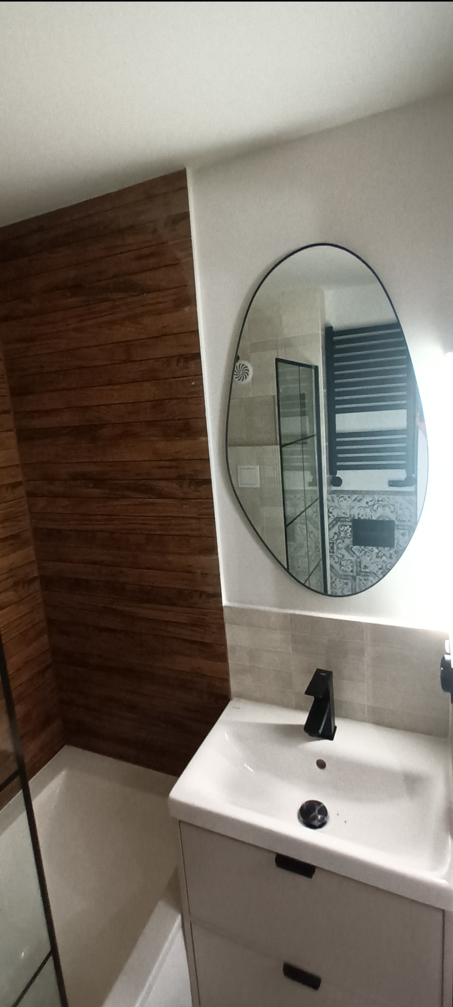 Nowoczesna łazienka po remoncie: drewnopodobna okładzina, asymetryczne lustro, biała umywalka z szafką i czarna bateria. Widoczny fragment kabiny prysznicowej.