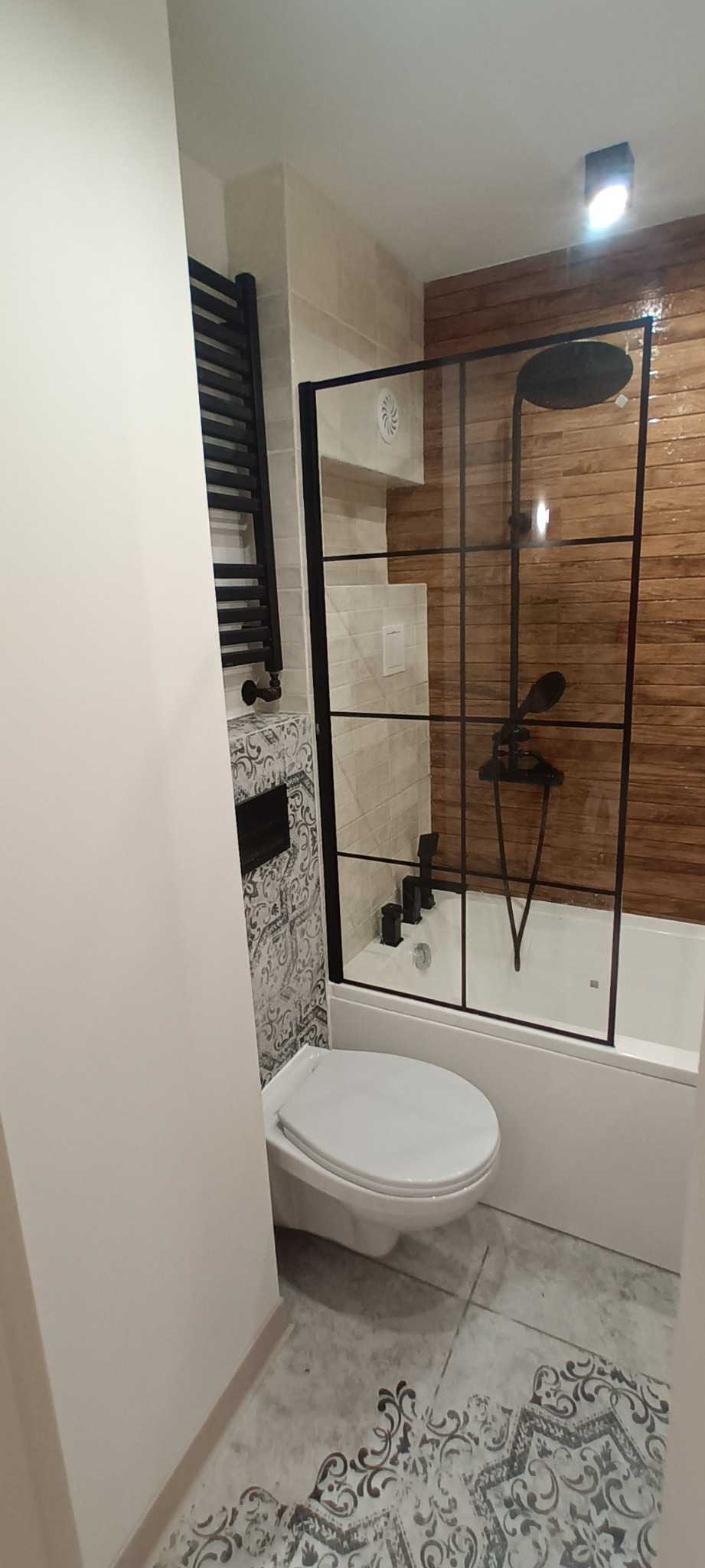 Nowoczesna łazienka po remoncie: kabina prysznicowa typu walk-in z czarnymi profilami, drewnopodobne płytki na ścianie, dekoracyjne płytki podłogowe i czarny grzejnik.