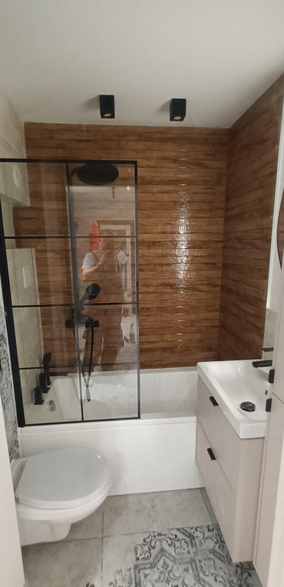 Wykończona łazienka z kabiną prysznicową typu walk-in z czarnymi profilami, drewnopodobnymi płytkami na ścianie i wzorzystą podłogą. Nowoczesne oświetlenie.